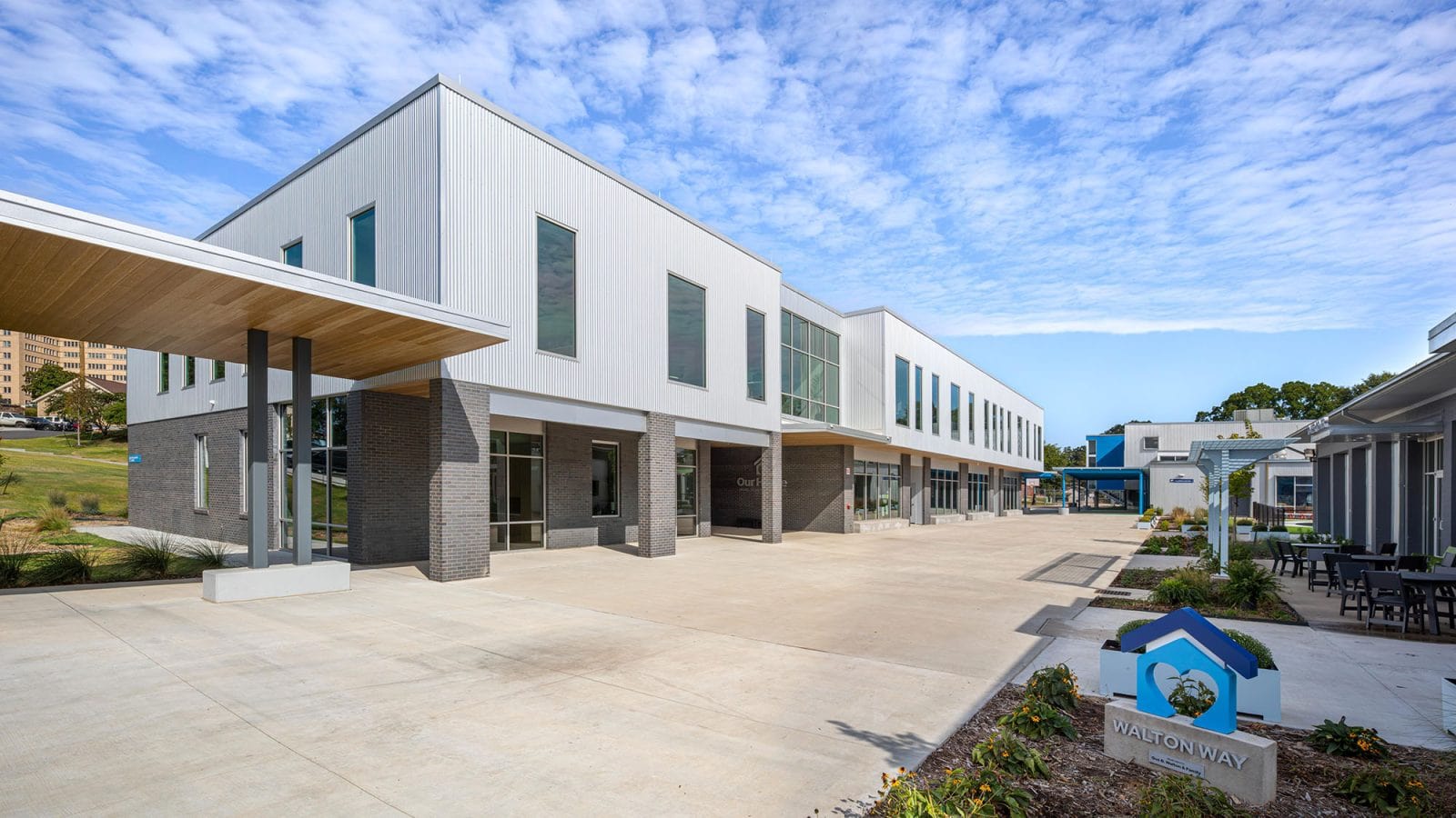 Modern two-story commercial building with large windows, flat roof, and a wide paved walkway; landscaped area and "WALTONWAY" sign in the foreground.