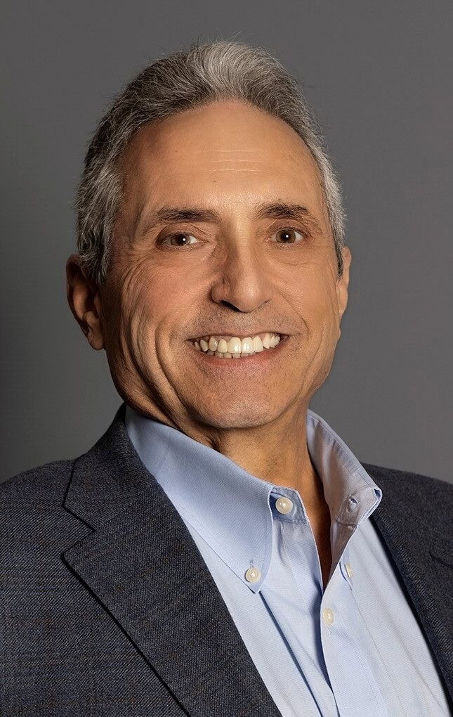 Smiling older man with gray hair wearing a blue dress shirt and dark blazer against a plain gray background.