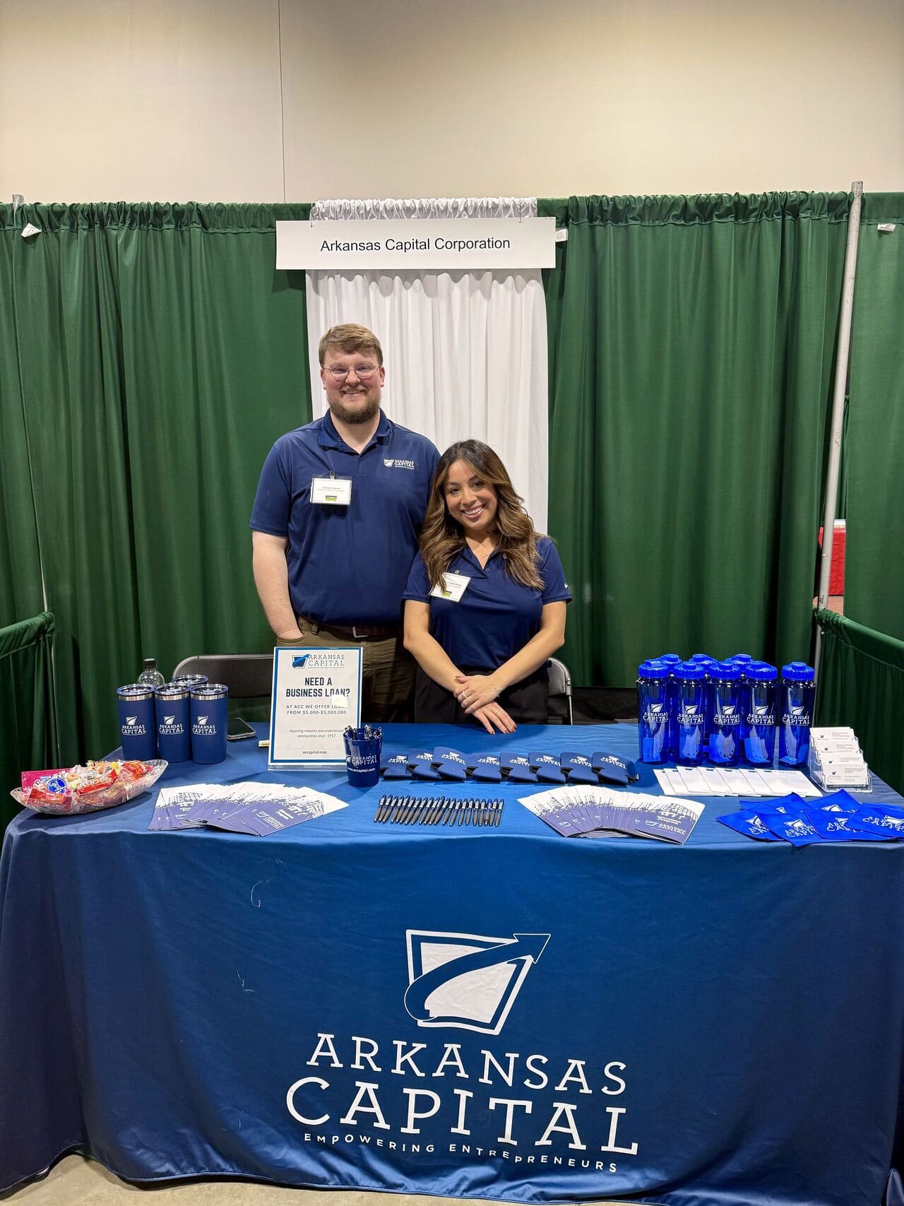 Two people stand behind a table covered with Arkansas Capital promotional items at an event booth, in front of a green curtain backdrop.