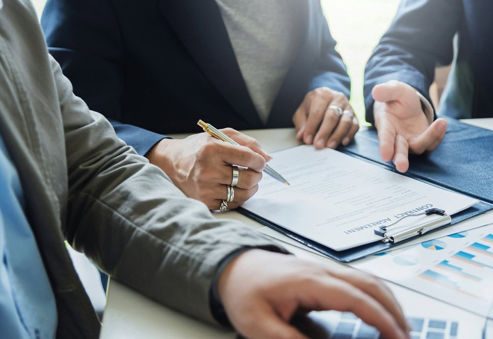 Three people sit at a table reviewing and signing a contract agreement, with charts and a laptop visible in the foreground.