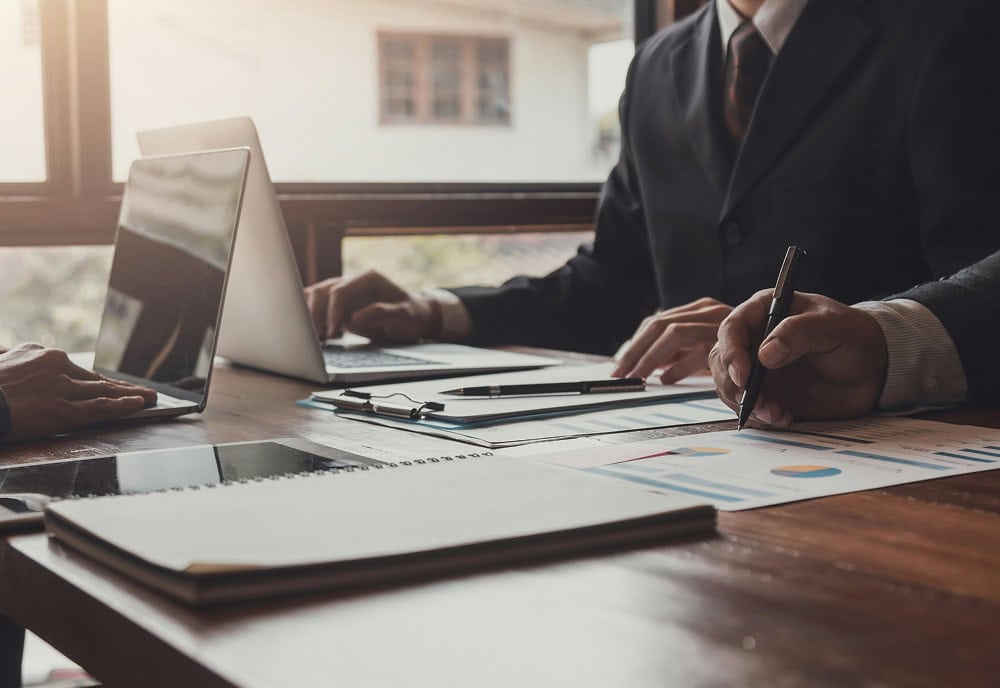 Two people in business attire work at a desk with a laptop, documents, charts, and a notebook, in a well-lit office setting.