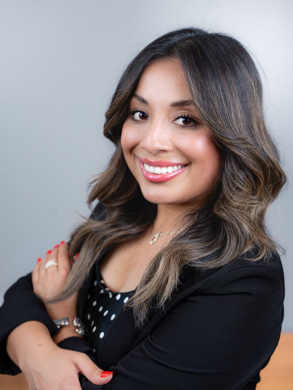 Woman with long wavy hair and a black blazer smiles at the camera with arms crossed, standing in front of a plain light gray background.