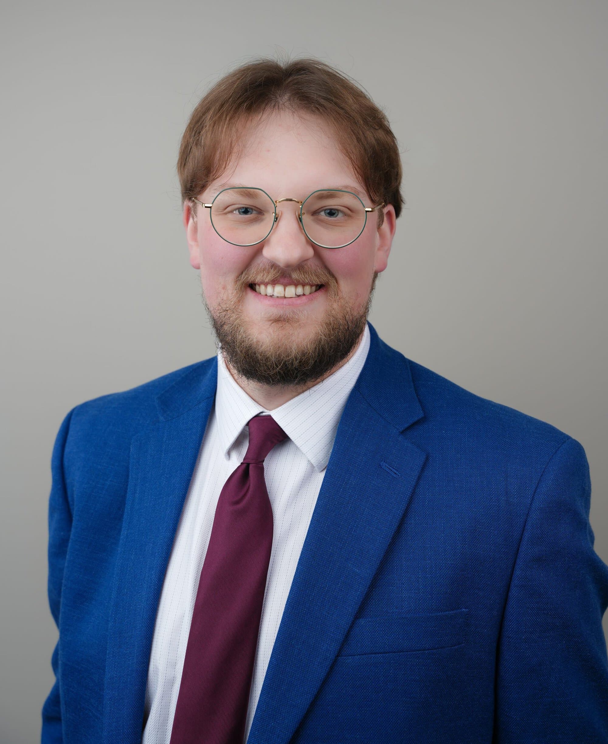 A man with glasses, a beard, and medium-length hair wearing a blue suit jacket, white shirt, and maroon tie, standing in front of a plain light background.