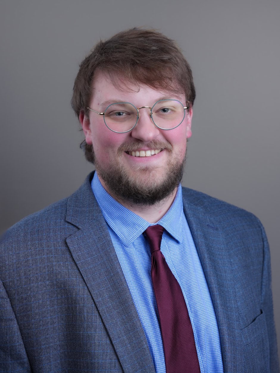 A man with glasses, a beard, and medium-length hair wearing a blue suit, light blue shirt, and maroon tie poses in front of a plain gray background.