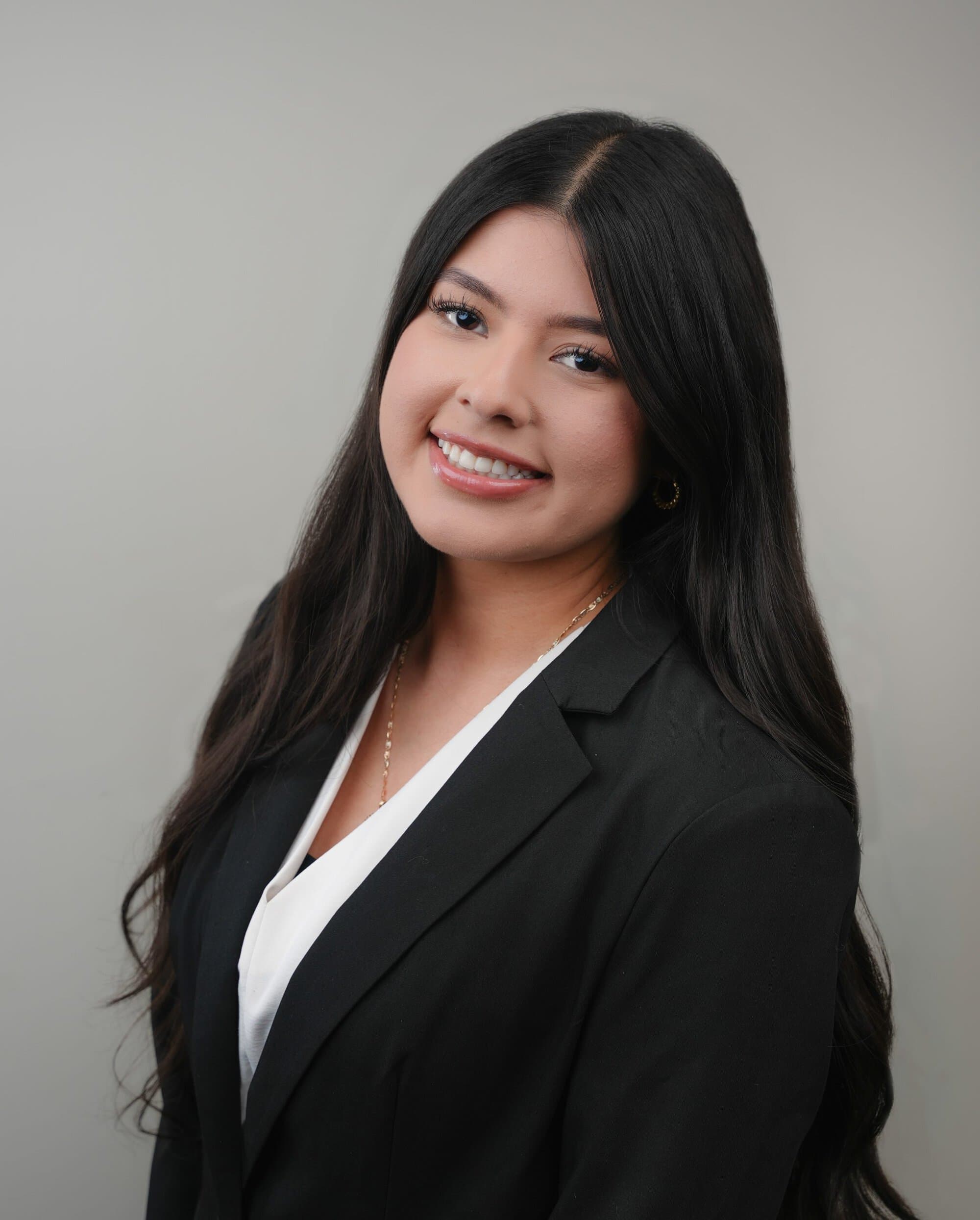 A woman with long dark hair wearing a black blazer and white blouse poses against a plain light gray background, smiling at the camera.