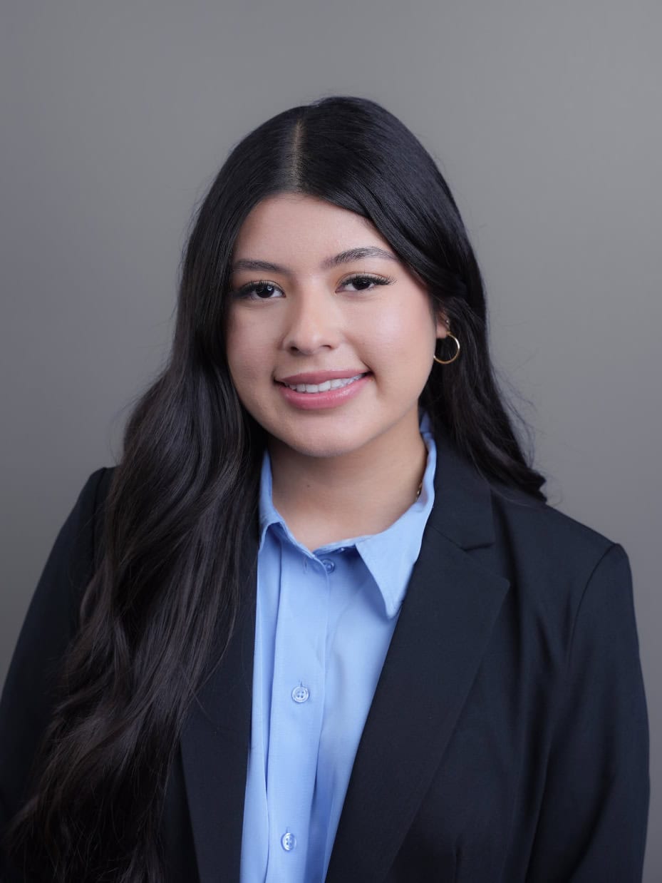 A young woman with long dark hair, wearing a black blazer and light blue shirt, poses against a plain gray background.