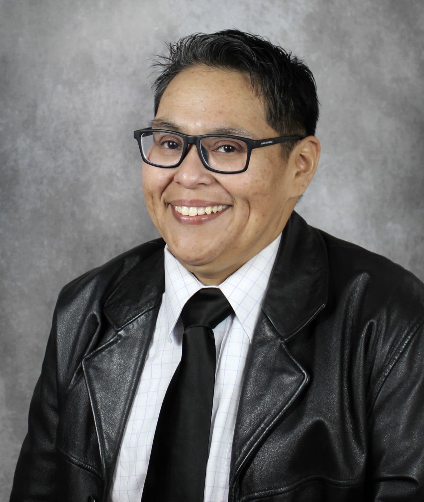 A person with short dark hair, glasses, and a leather jacket over a white shirt and tie, smiling against a gray studio background.