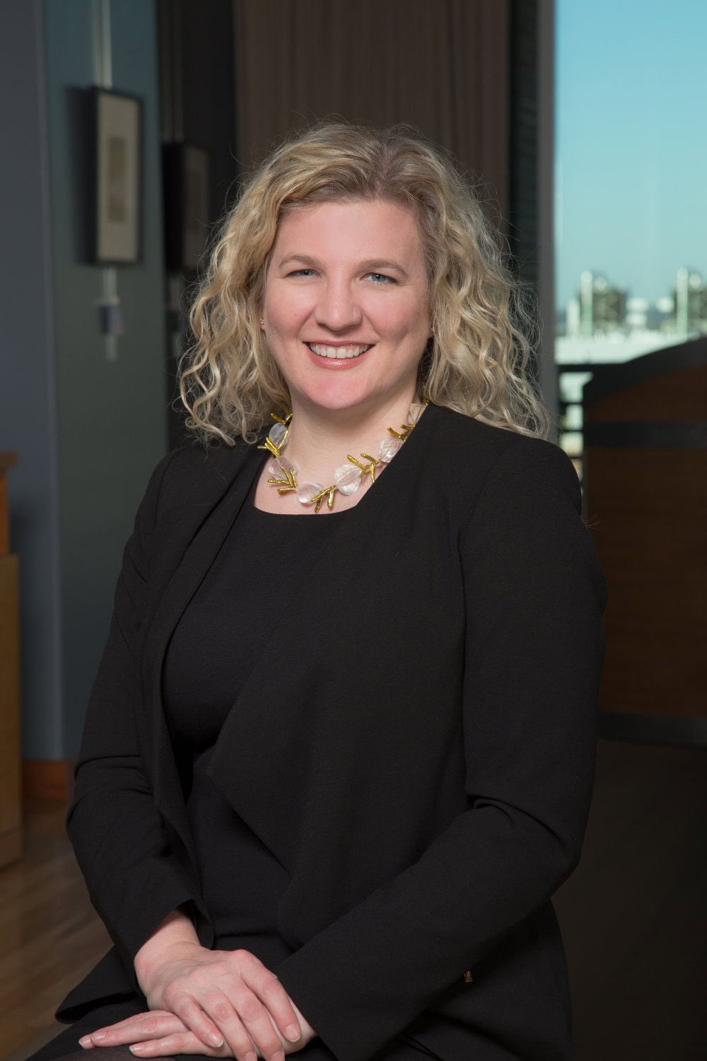 A woman with curly blonde hair wearing a black blazer and necklace sits and smiles at the camera in an indoor setting.