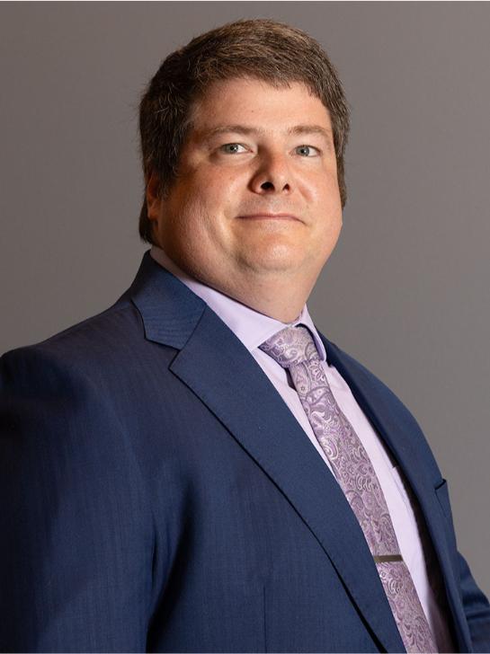 A man in a blue suit, lavender dress shirt, and patterned tie poses in front of a plain gray background.
