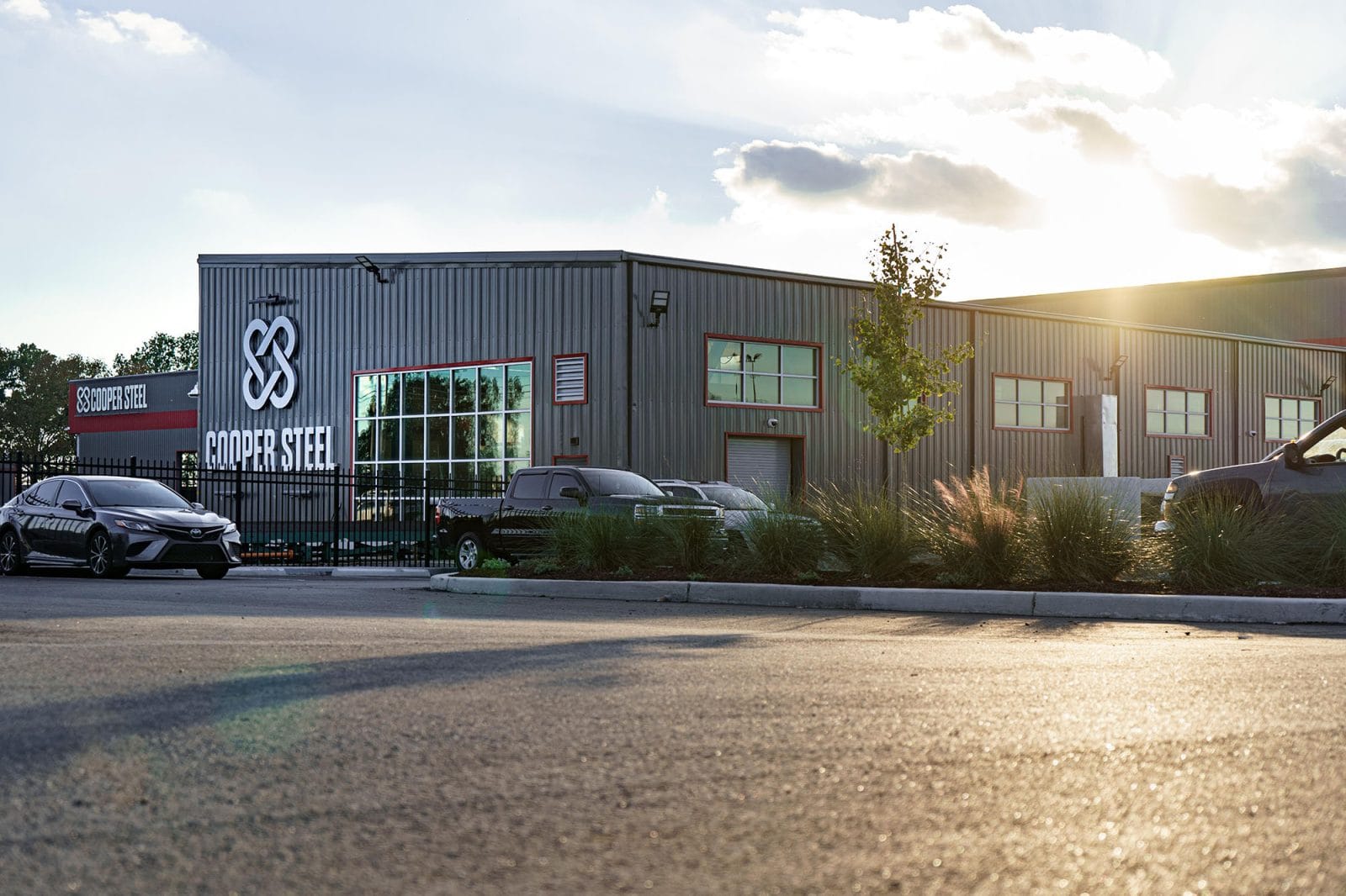 Exterior view of the Cooper Steel building during daylight, with parked cars and a police car in front, and the sun shining in the background.