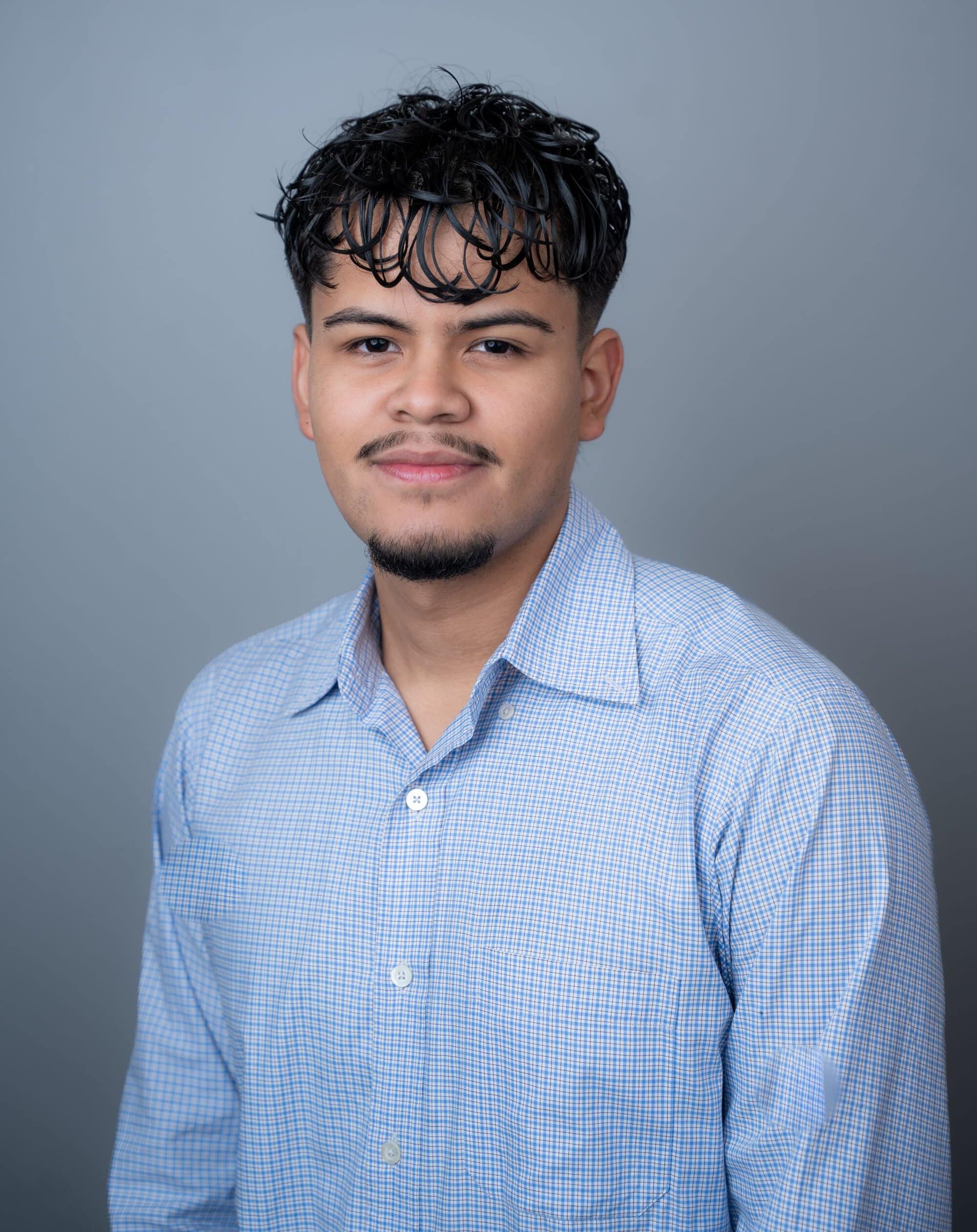 A young man with dark, textured hair and a goatee, wearing a light blue checkered button-down shirt.
