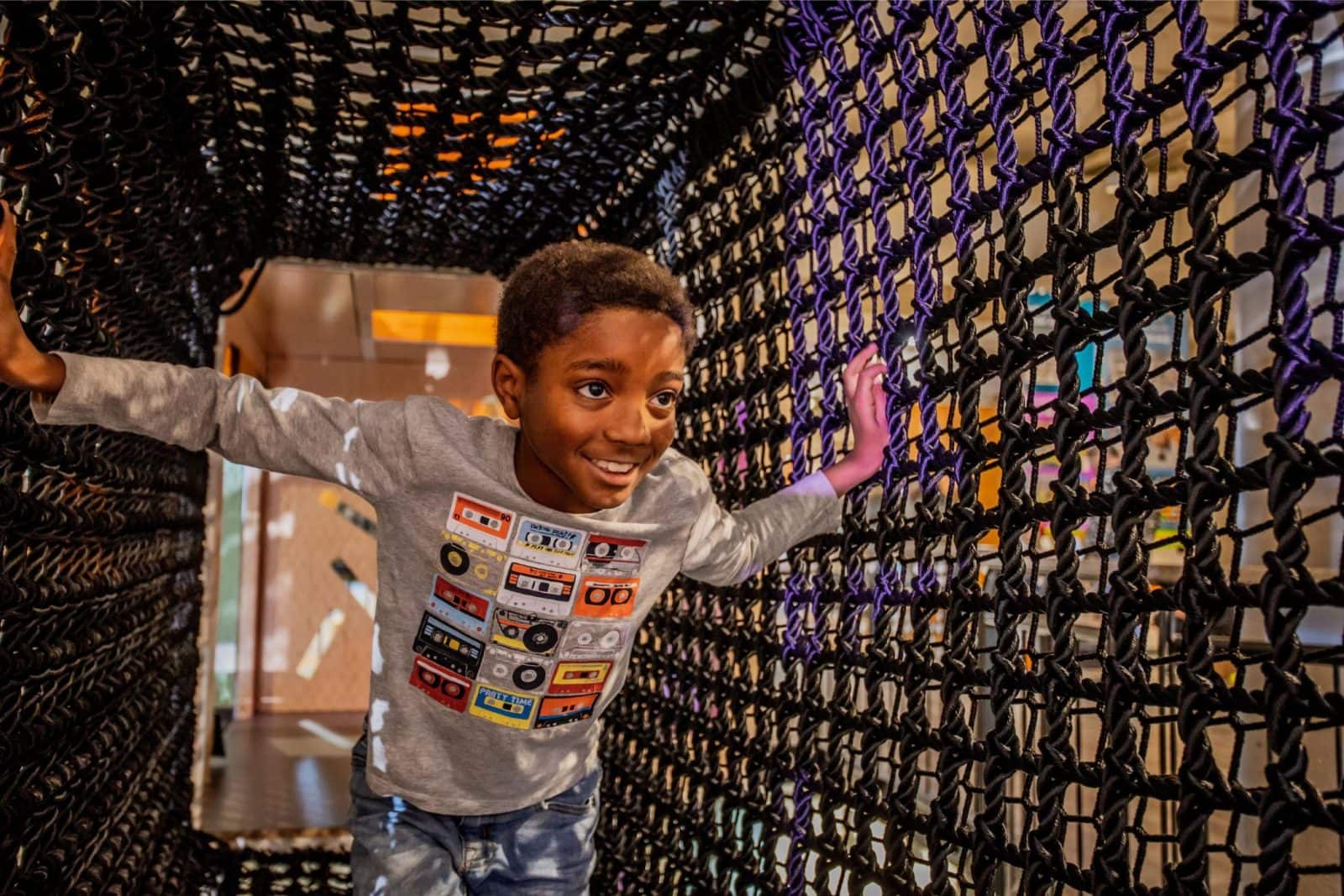 A child wearing a gray shirt with a cassette tape design crawls through a black rope tunnel in an indoor play area.