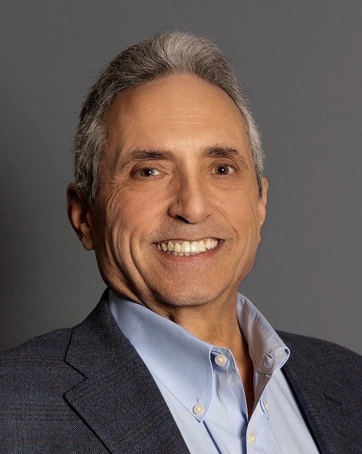 A middle-aged man with gray hair wearing a blue dress shirt and a dark blazer, smiling in front of a plain gray background.