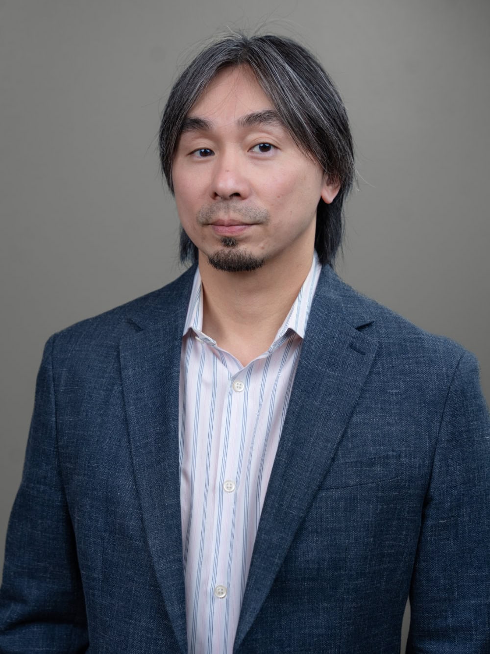 A man with straight, shoulder-length gray hair and a goatee wears a blue blazer over a striped shirt, standing against a plain gray background.