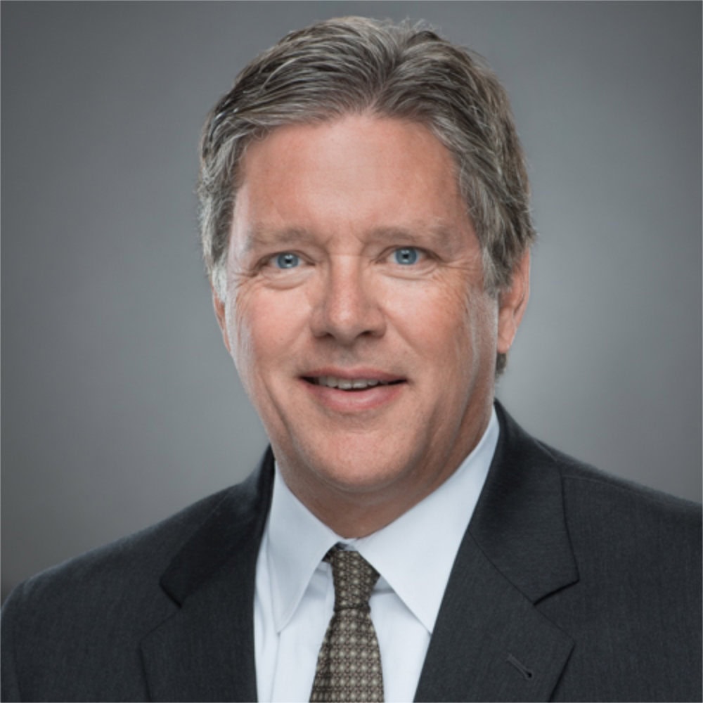 A middle-aged man with grayish-brown hair wearing a dark suit, white shirt, and patterned tie poses against a neutral gray background.