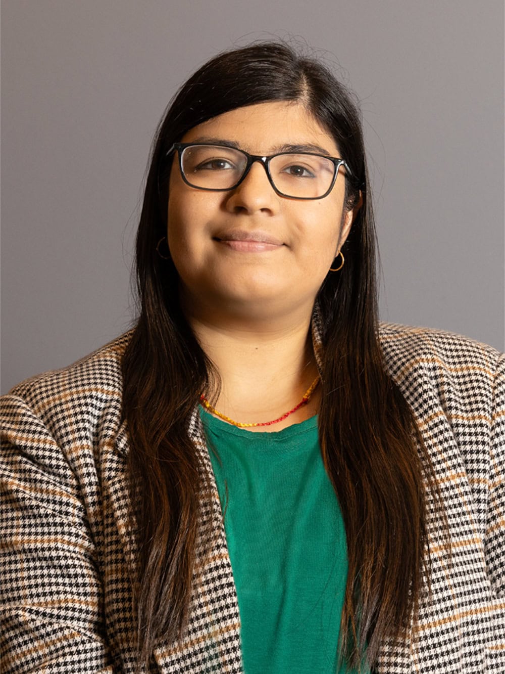 A person with long dark hair, glasses, and a plaid blazer over a green shirt, facing the camera with a neutral expression against a plain gray background.