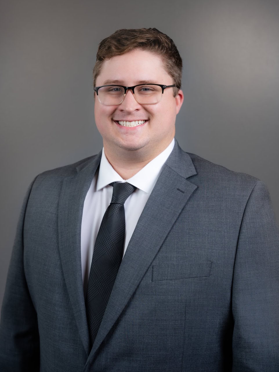 A man wearing glasses and a gray suit with a white shirt and black tie stands smiling against a plain gray background.