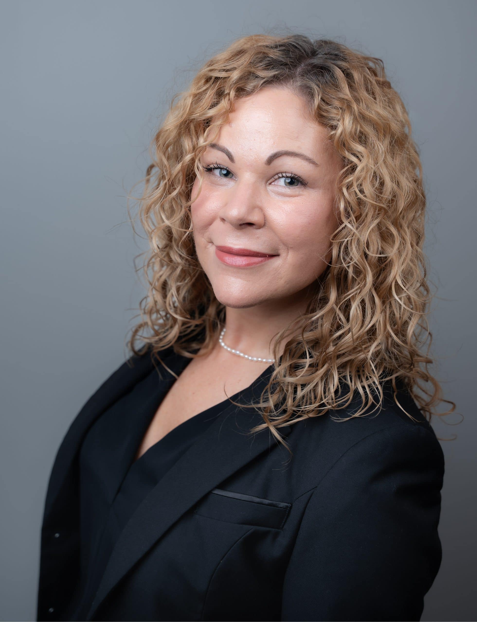 Headshot of a woman with curly blonde hair wearing a black blazer and a pearl necklace.