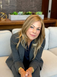 A woman with blonde hair wearing a gray suit sits on a light-colored sofa, looking at the camera, with a planter and table in the background.