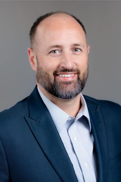 A man with a trimmed beard and short hair, wearing a navy blazer and checked shirt, poses against a plain gray background.