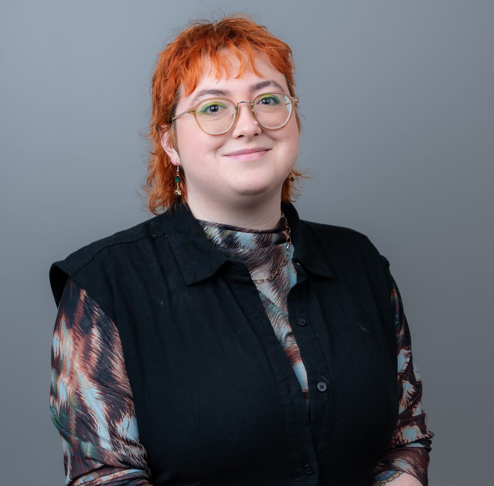 A person with short, orange hair and round glasses smiles against a gray background, wearing a patterned top under a black vest.