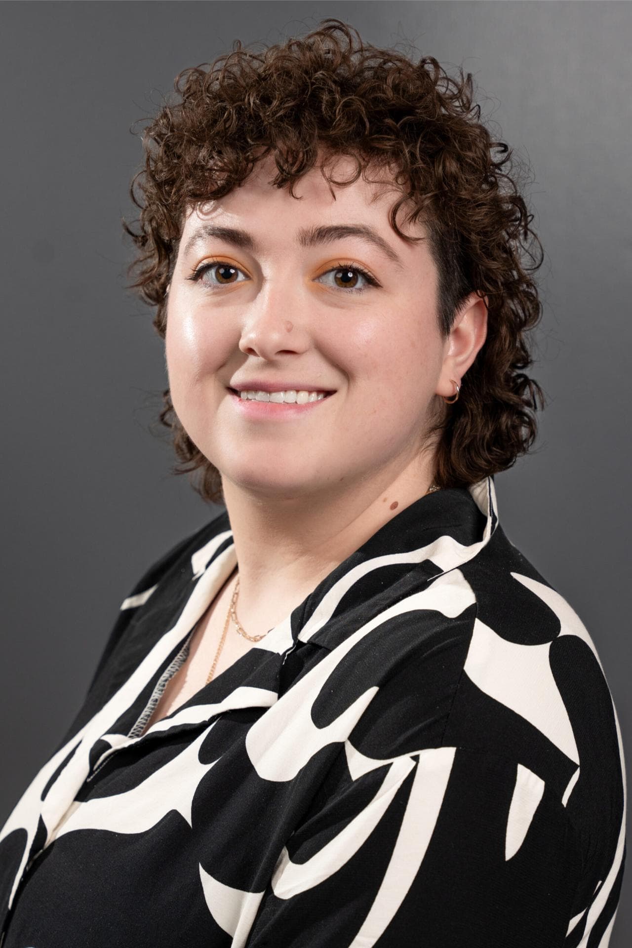 A person with short, curly brown hair and light skin smiles at the camera, wearing a black and white patterned shirt, against a plain grey background.