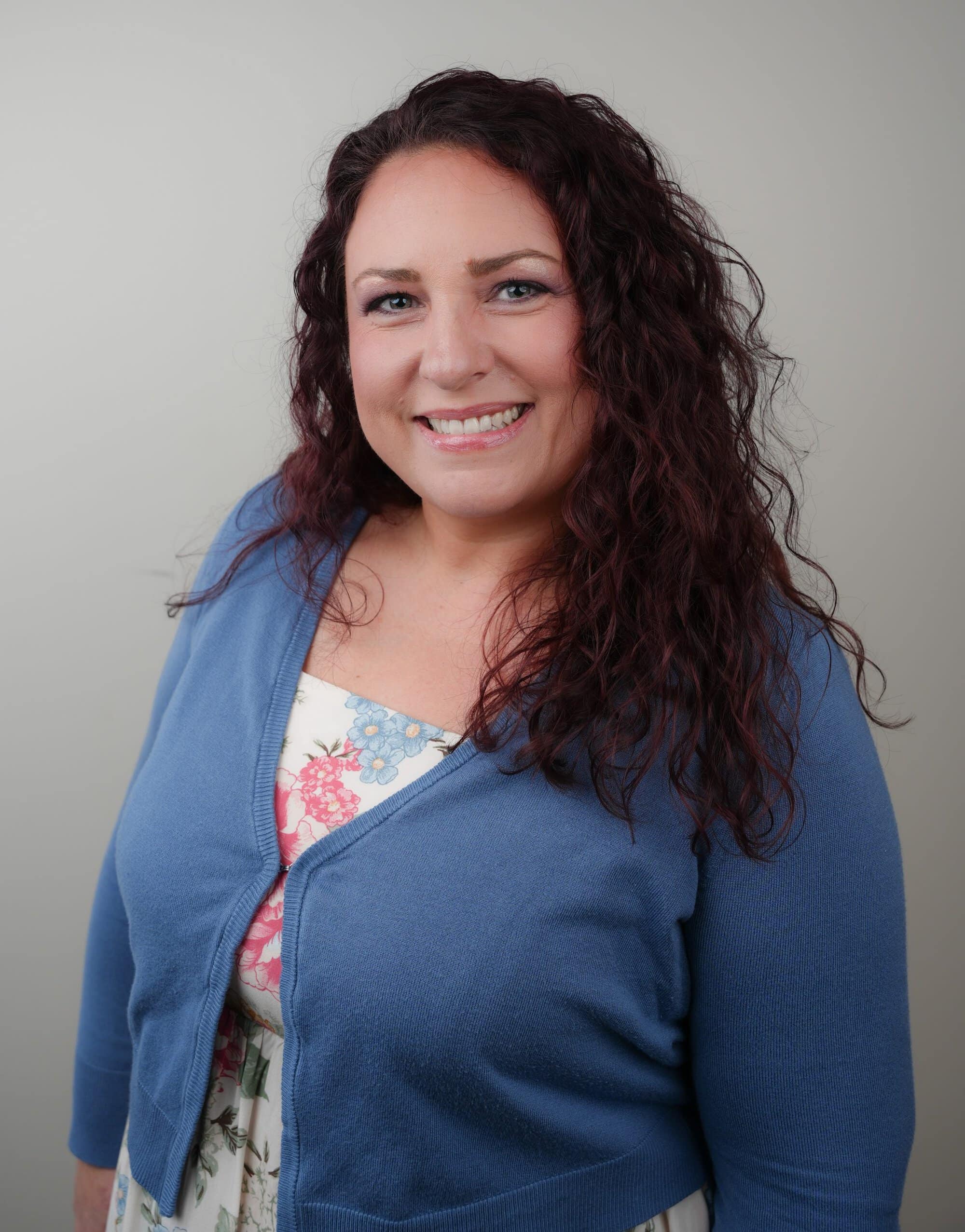 A woman with long curly brown hair wearing a blue cardigan over a floral dress smiles at the camera against a plain background.