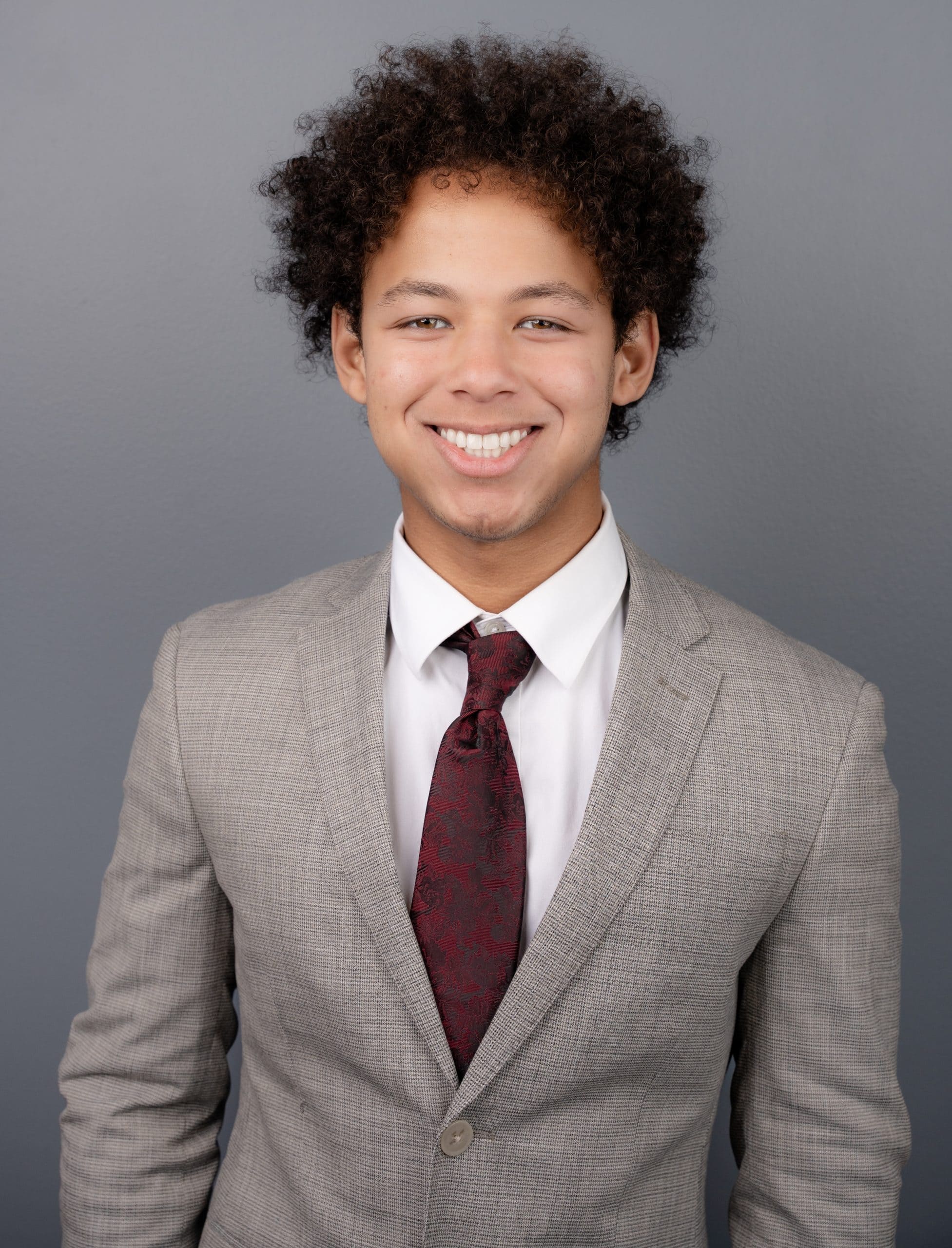A person with curly hair wearing a light gray suit, white shirt, and maroon tie, smiles while standing against a plain gray background.