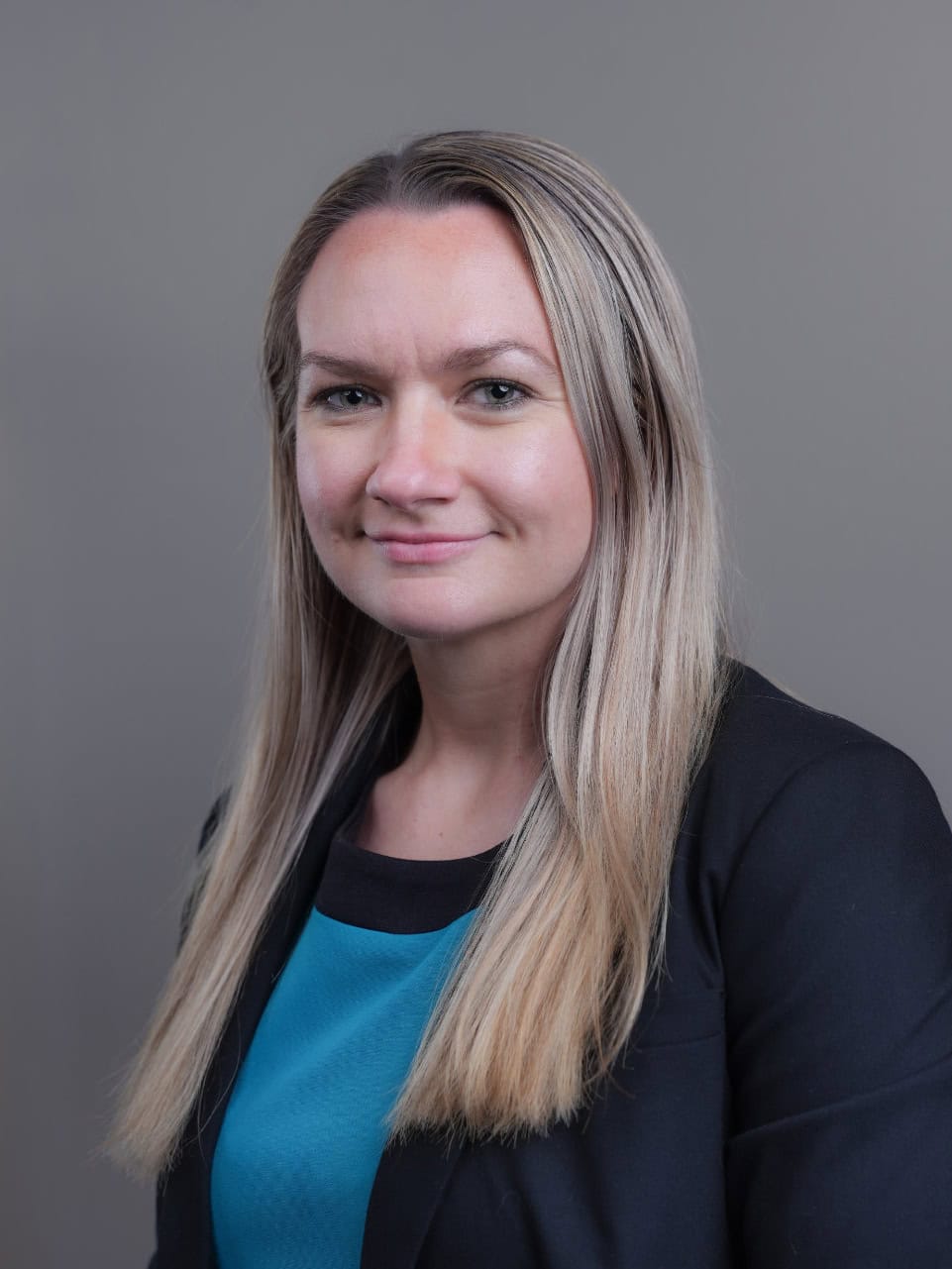 A woman with straight, long blonde hair wearing a black blazer and teal top poses in front of a plain gray background.