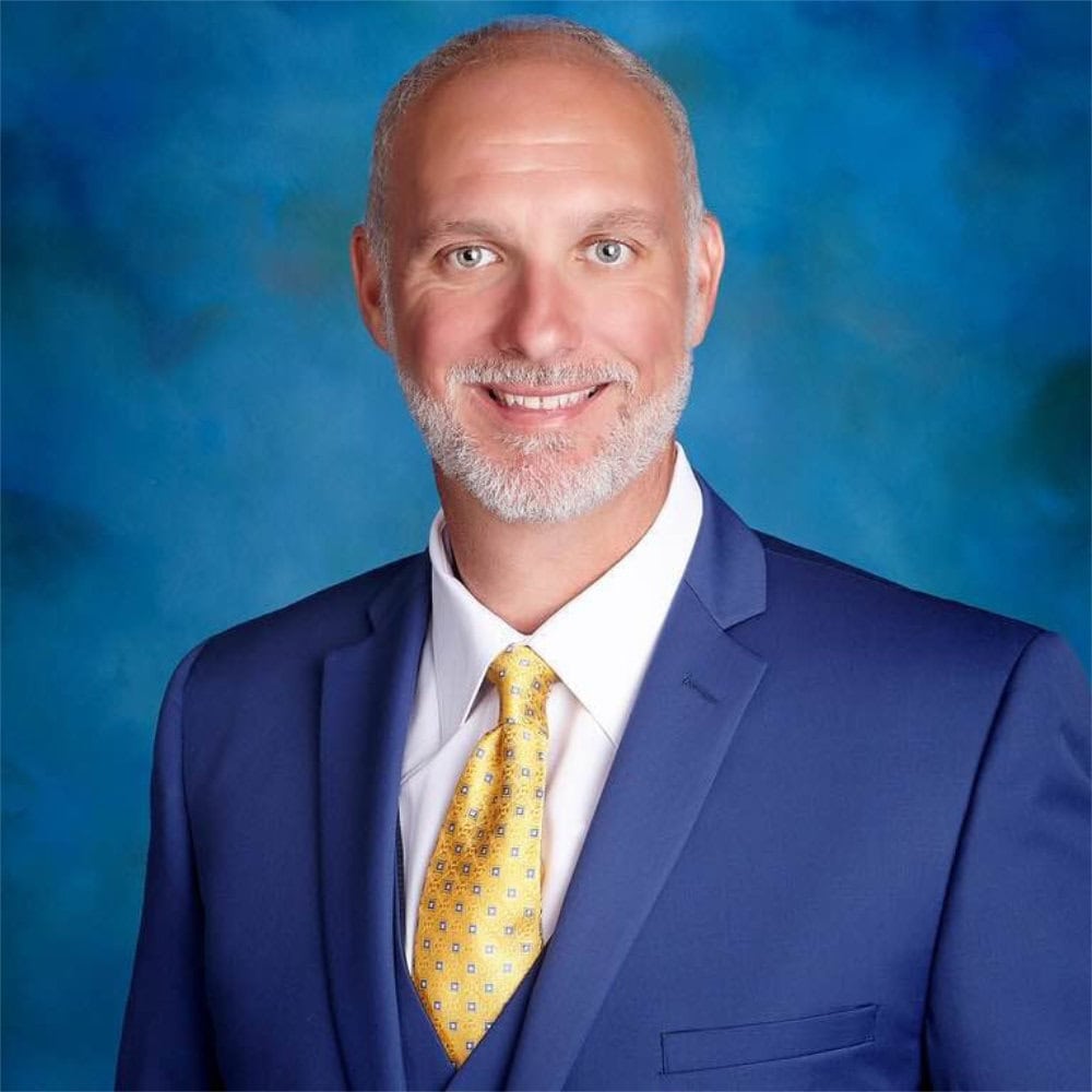 A man in a blue suit, white shirt, and yellow tie poses in front of a blue studio backdrop, smiling at the camera.
