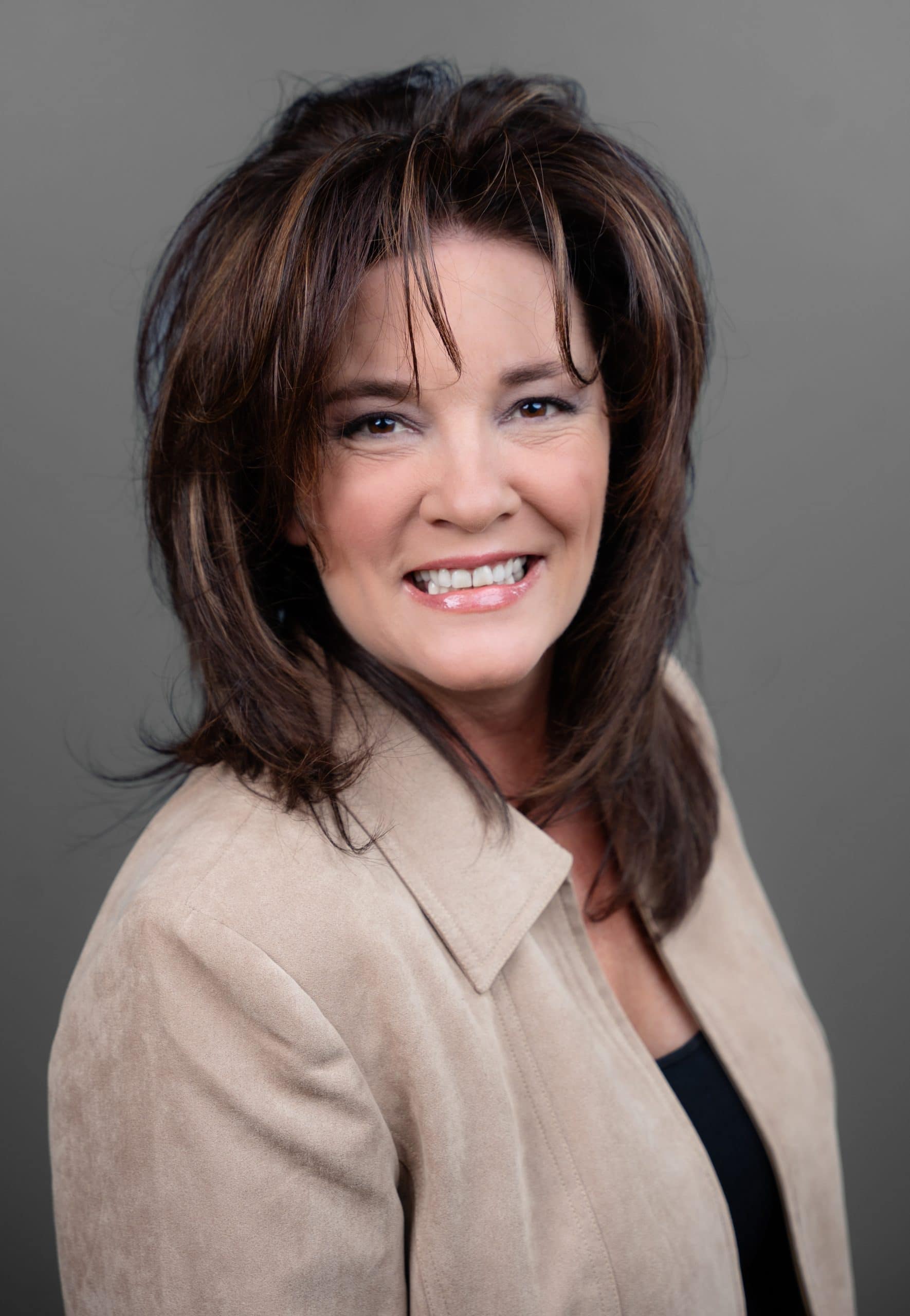 Woman with shoulder-length brown hair wearing a beige jacket and a black top, smiling at the camera against a gray background.