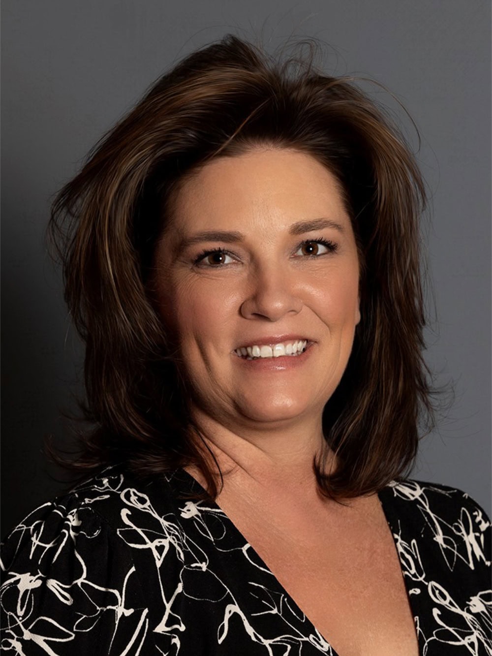 Woman with shoulder-length brown hair and a patterned black and white top smiles in front of a plain gray background.