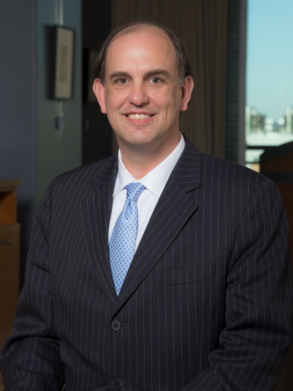 A man in a dark pinstripe suit and light blue tie smiles at the camera in an office setting.