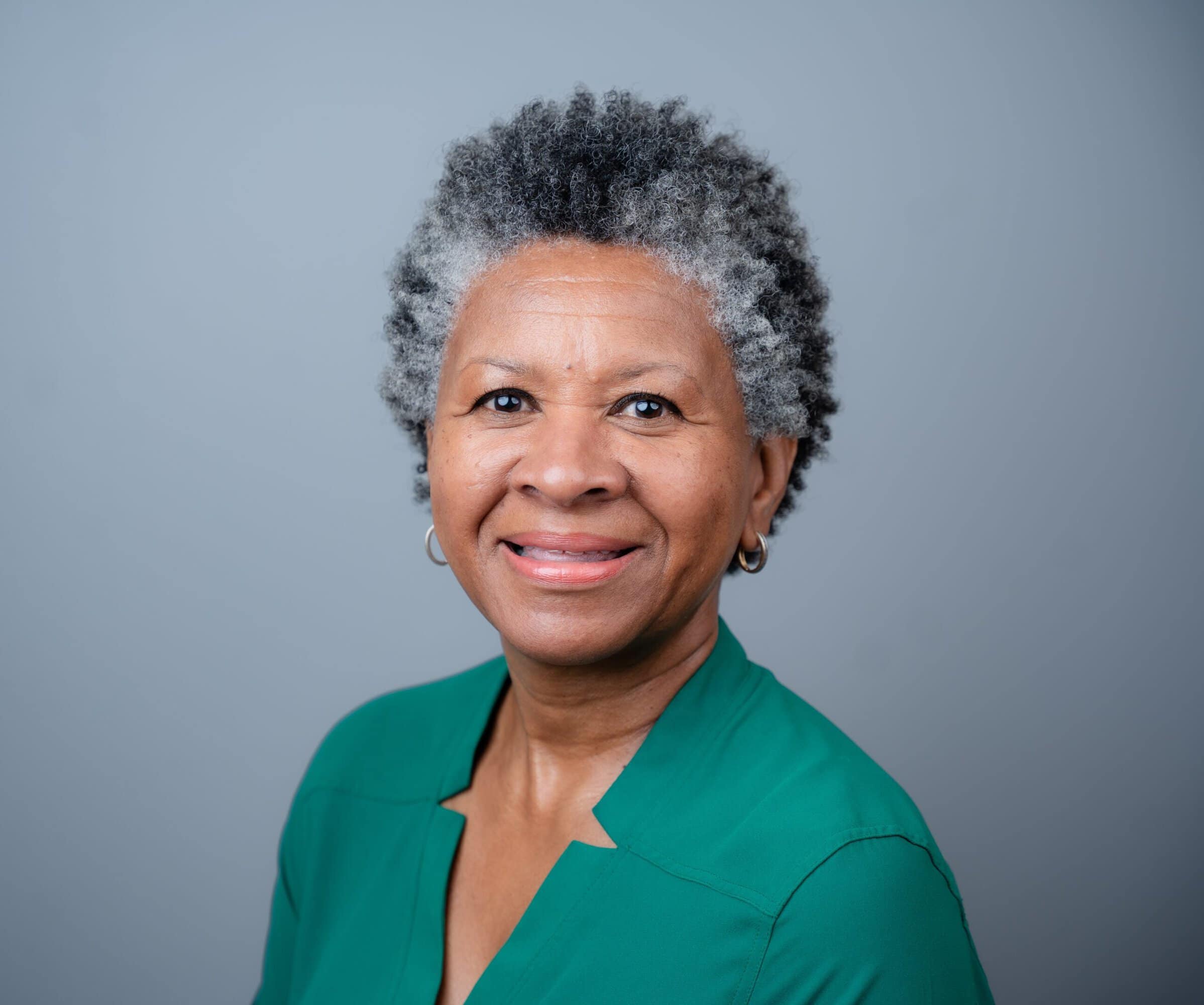 A smiling middle-aged woman with short, gray, curly hair wearing a green top.