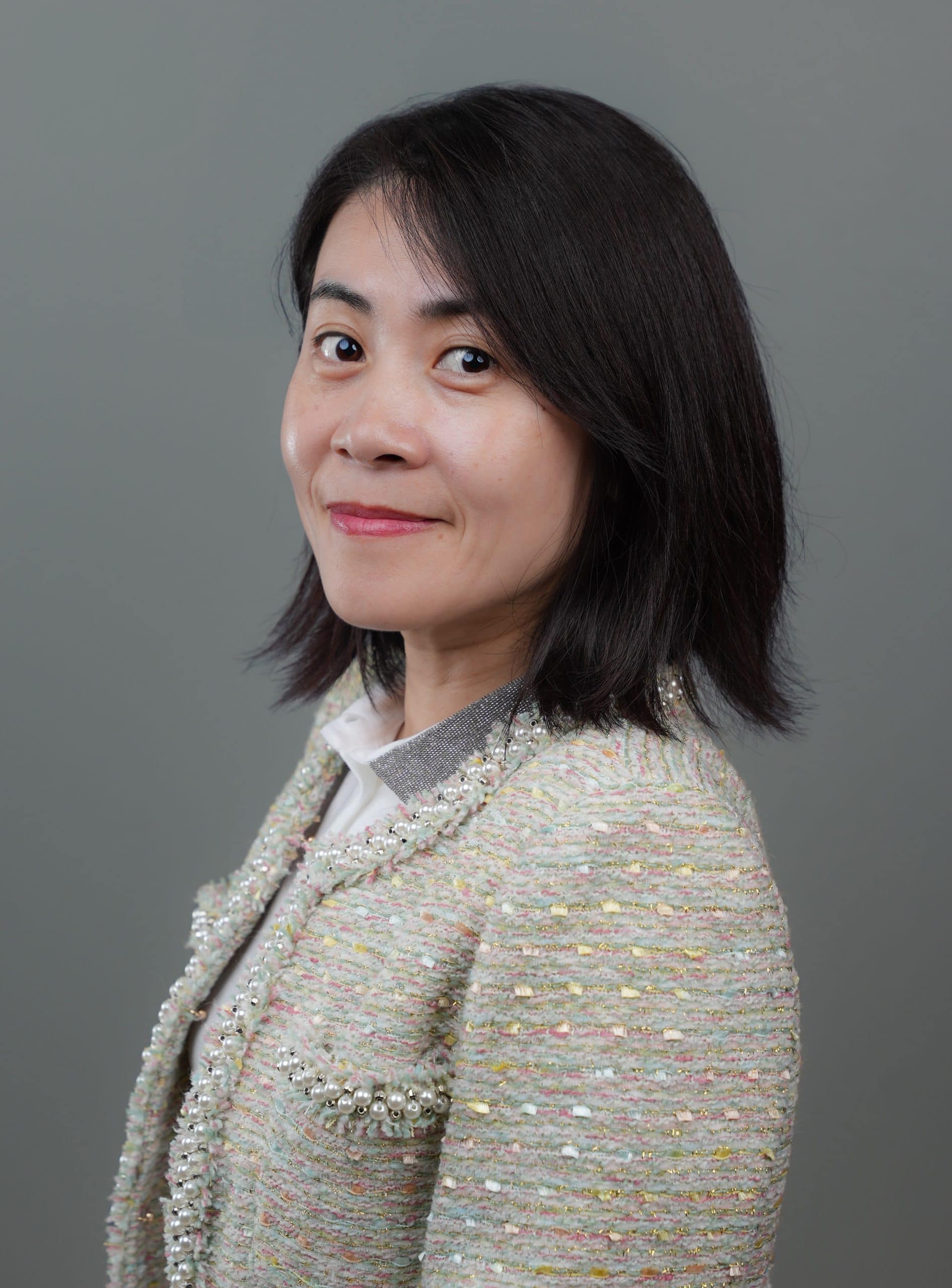 Woman with medium-length dark hair wearing a textured light-colored blazer, posing in profile against a plain gray background.