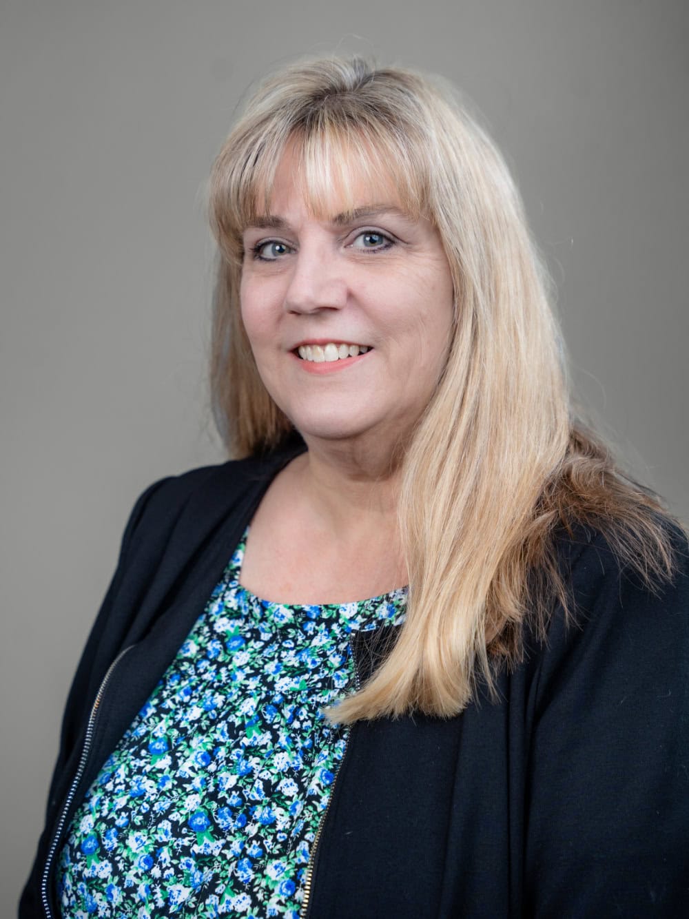 A woman with long blonde hair wearing a black jacket and a blue floral top poses for a professional portrait against a plain gray background.