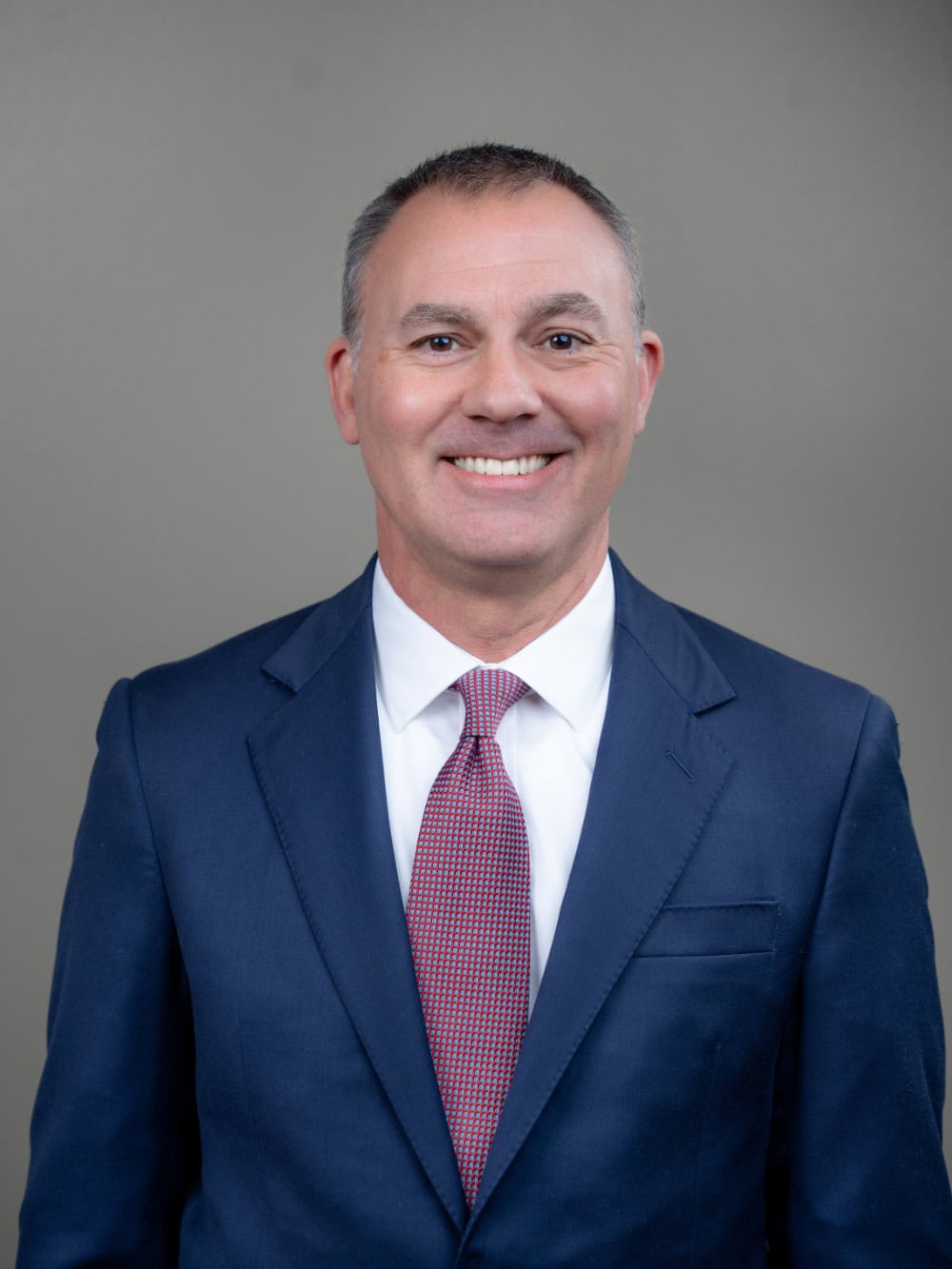 A man in a blue suit, white shirt, and red patterned tie stands facing the camera and smiles against a plain gray background.