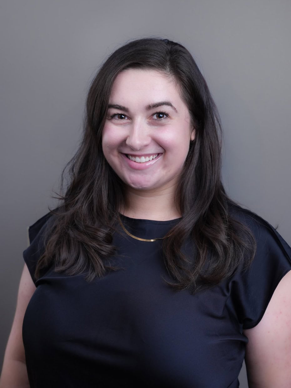 A woman with long dark hair, wearing a black top and a necklace, smiles at the camera against a plain gray background.