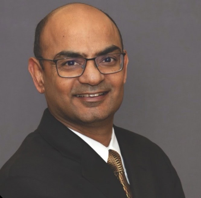 A man with glasses wearing a dark suit, white shirt, and gold tie, smiling in front of a plain grey background.