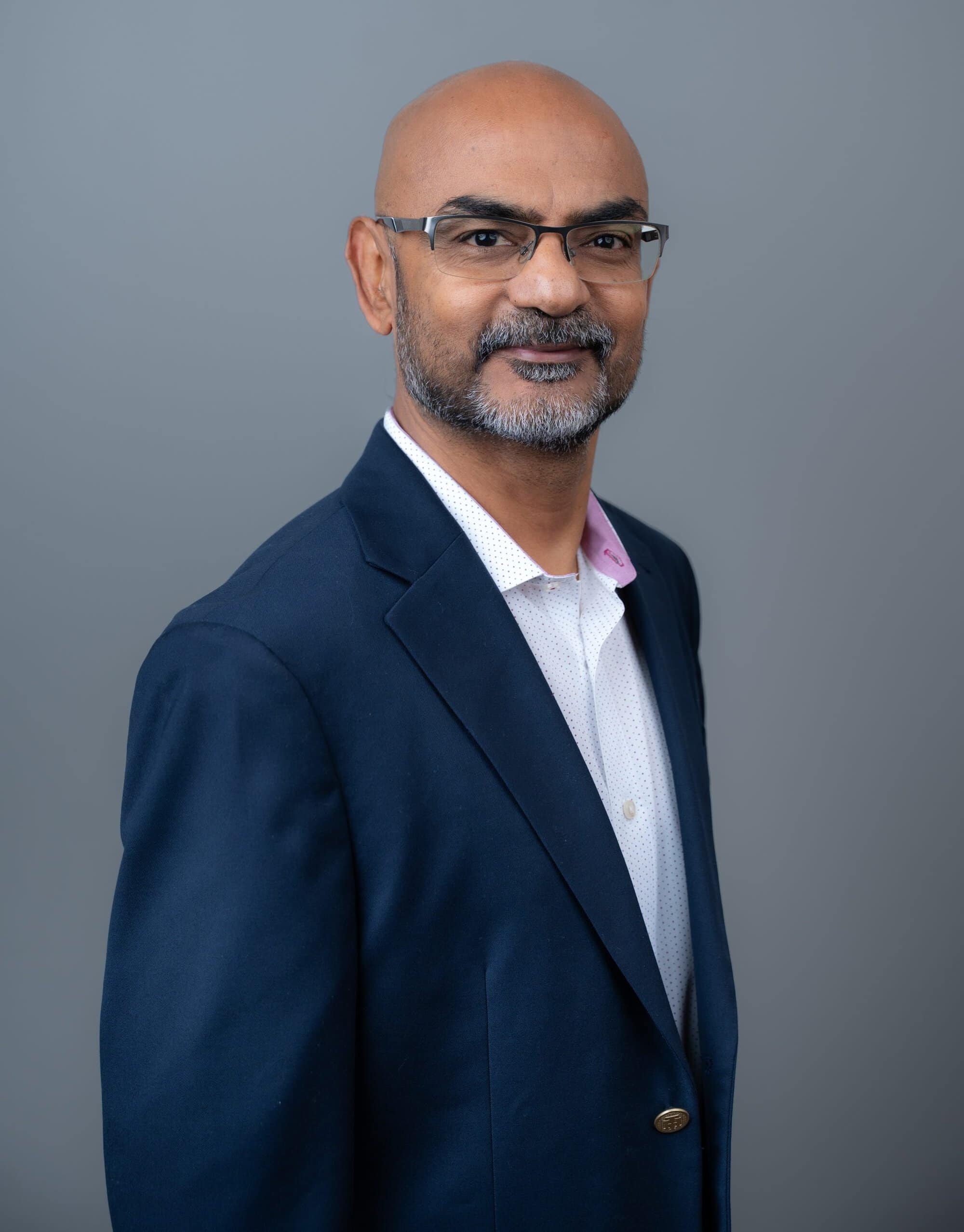 A man with a shaved head and a beard wearing glasses, a white collared shirt, and a navy blue blazer.
