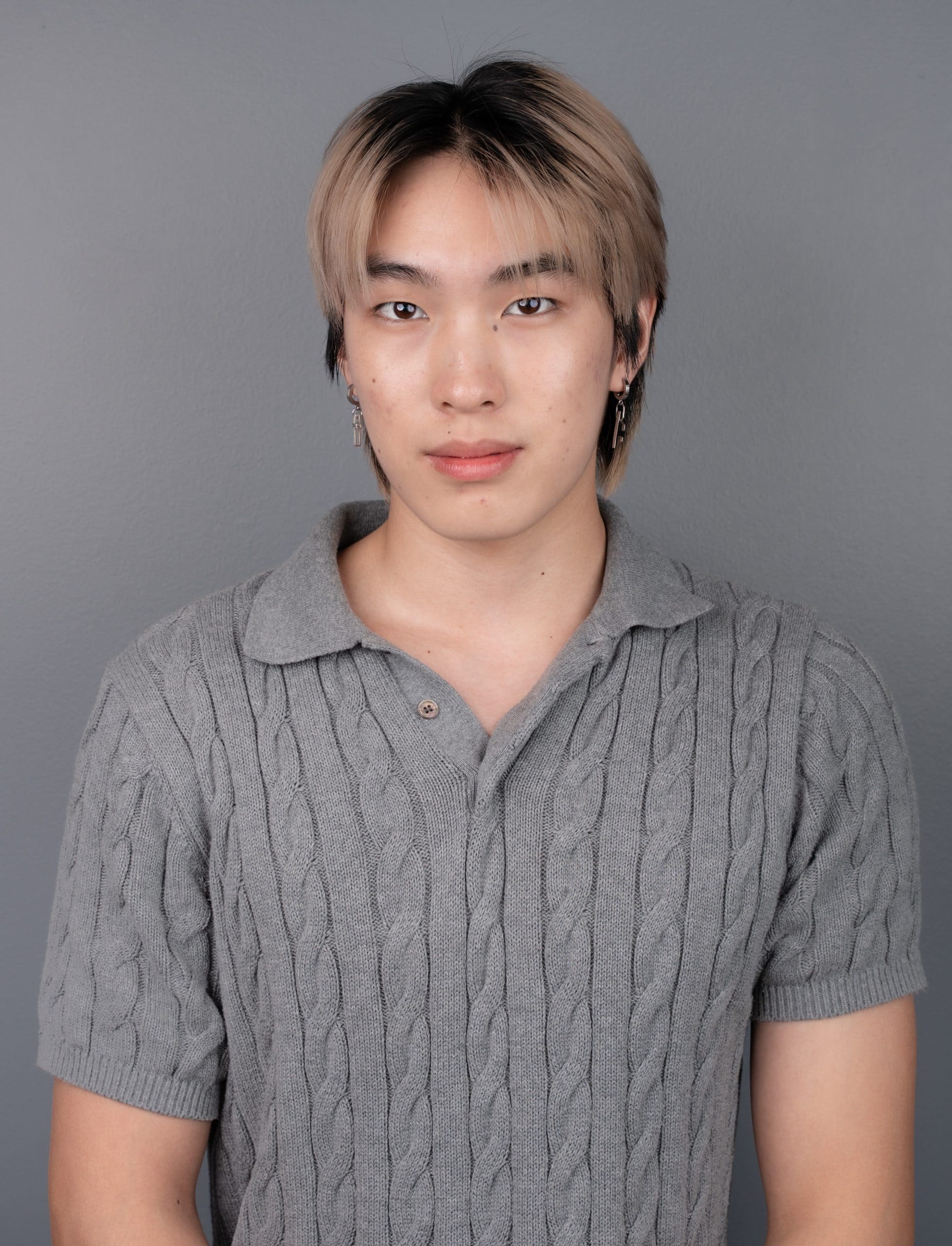 A person with straight, chin-length hair wearing a grey cable-knit short-sleeve shirt stands against a plain grey background, looking at the camera with a neutral expression.