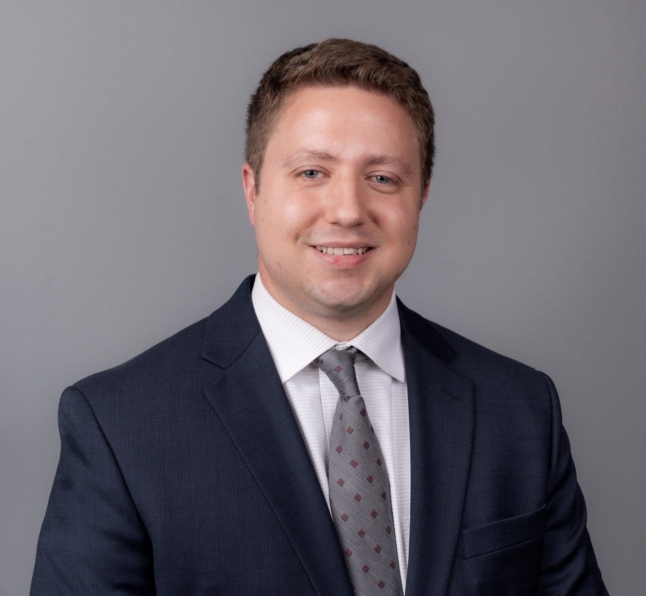 A man in a dark suit, white shirt, and patterned tie poses against a plain gray background, smiling slightly at the camera.