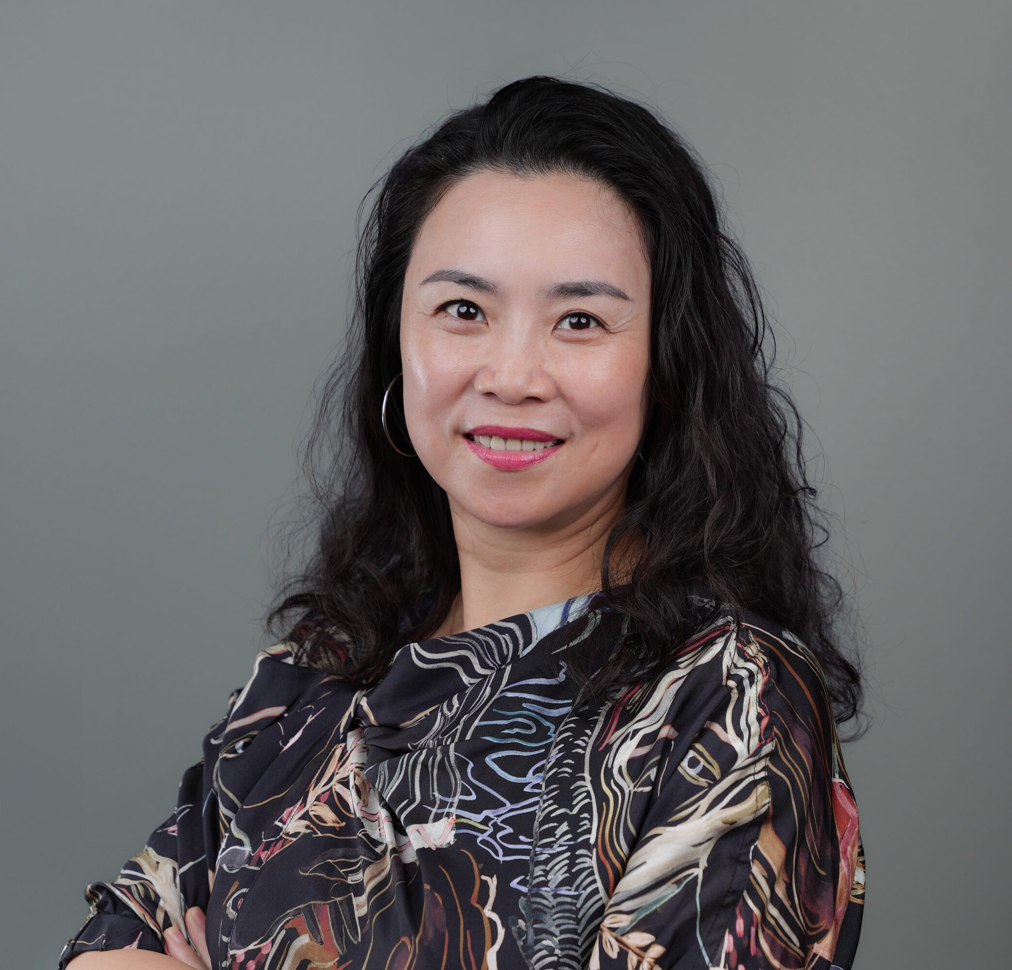 A woman with long curly dark hair, wearing a patterned top, hoop earrings, and pink lipstick, smiles at the camera against a plain grey background.