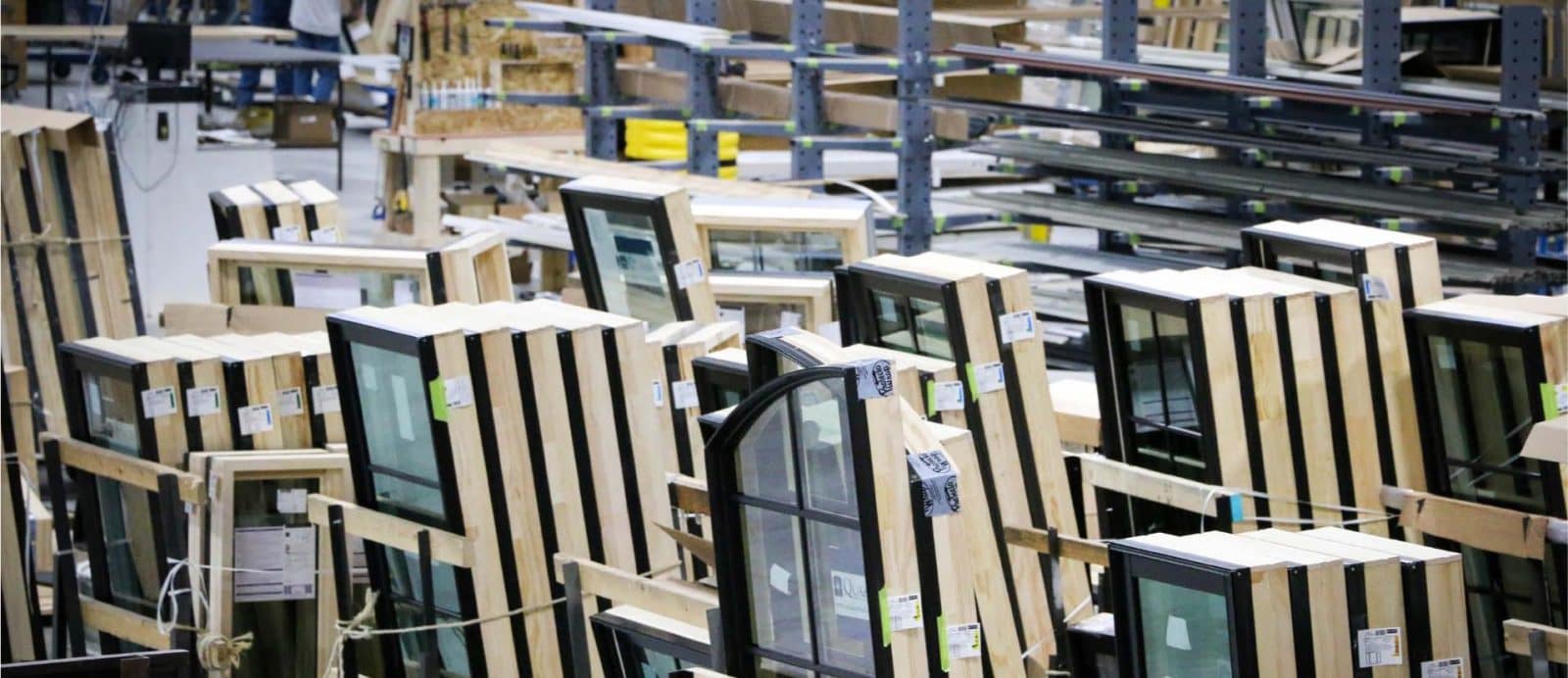 Factory interior with multiple framed glass windows organized in rows, surrounded by metal shelves and other building materials.