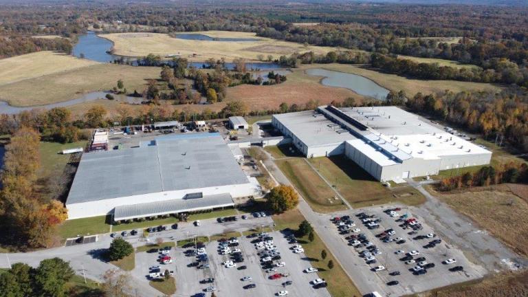 Aerial view of a large industrial facility with two main buildings, adjacent parking lot with many cars, and surrounding fields, trees, and ponds.