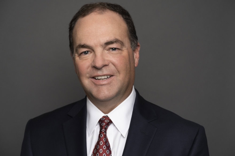 A man in a dark suit, white shirt, and patterned tie smiles in front of a plain gray background.