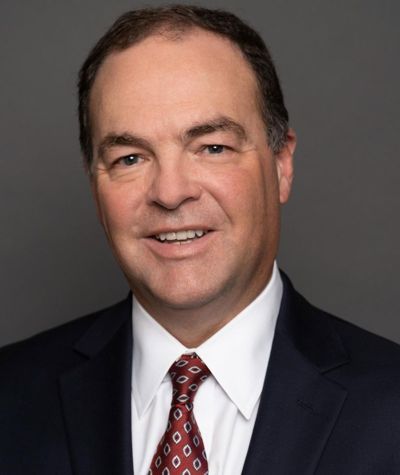 A middle-aged man in a dark suit, white shirt, and patterned tie poses against a plain gray background.