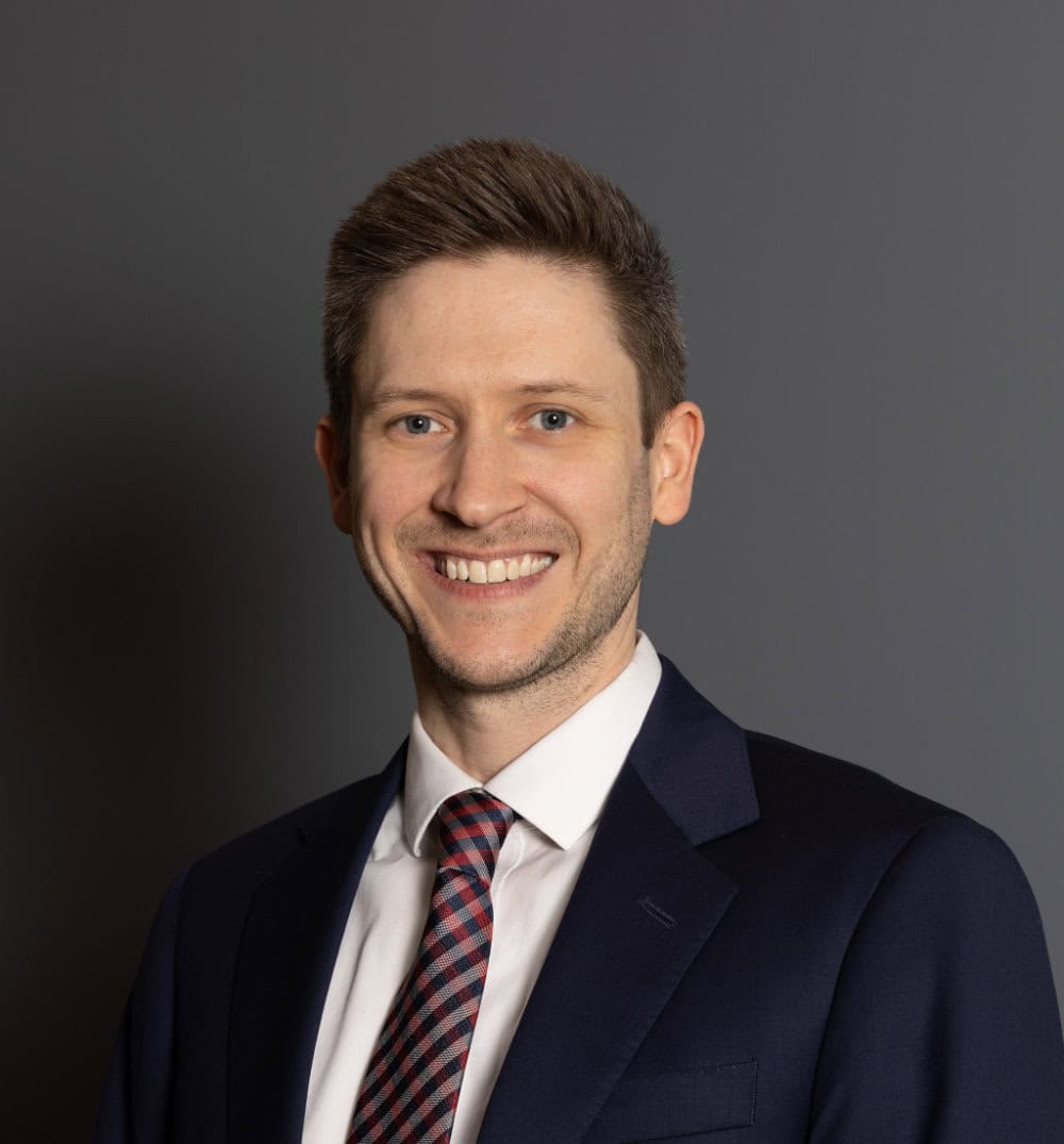 A man in a dark suit, white shirt, and patterned tie stands in front of a plain gray background, smiling at the camera.