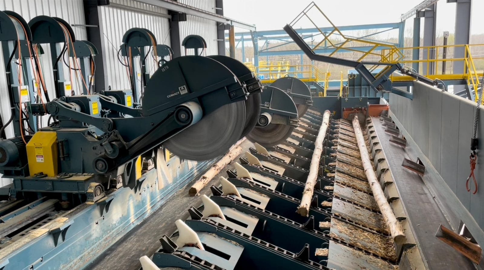 Large circular saws cut logs on an automated conveyor system inside an industrial sawmill.