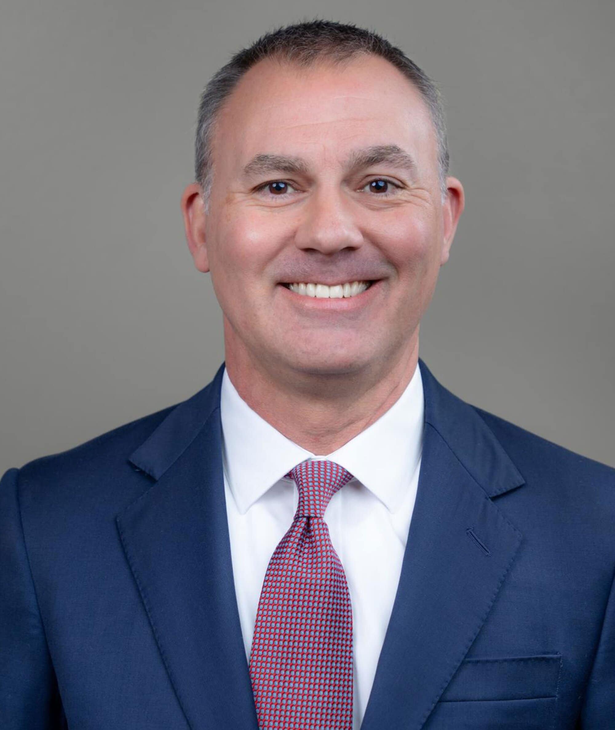 A man in a blue suit, white shirt, and patterned red tie smiling at the camera, posed against a plain gray background.